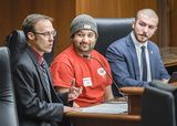 Matt Toburen sits at the testifiers' table and speaks to someone out of view. Cree Gordon and Representative Cantrell sit next to him and watch as he speaks.